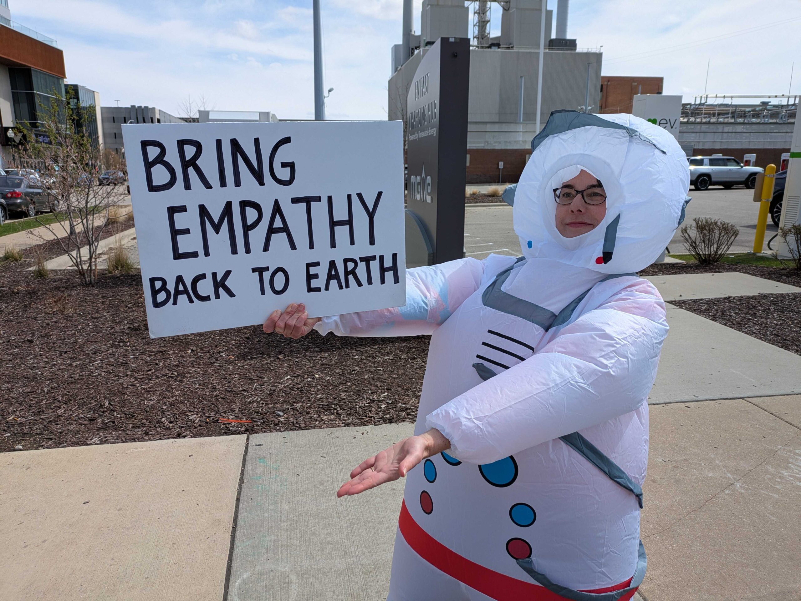 A person in an inflatable astronaut costume holds a sign that reads Bring Empathy Back to Earth on a sidewalk in an urban area.