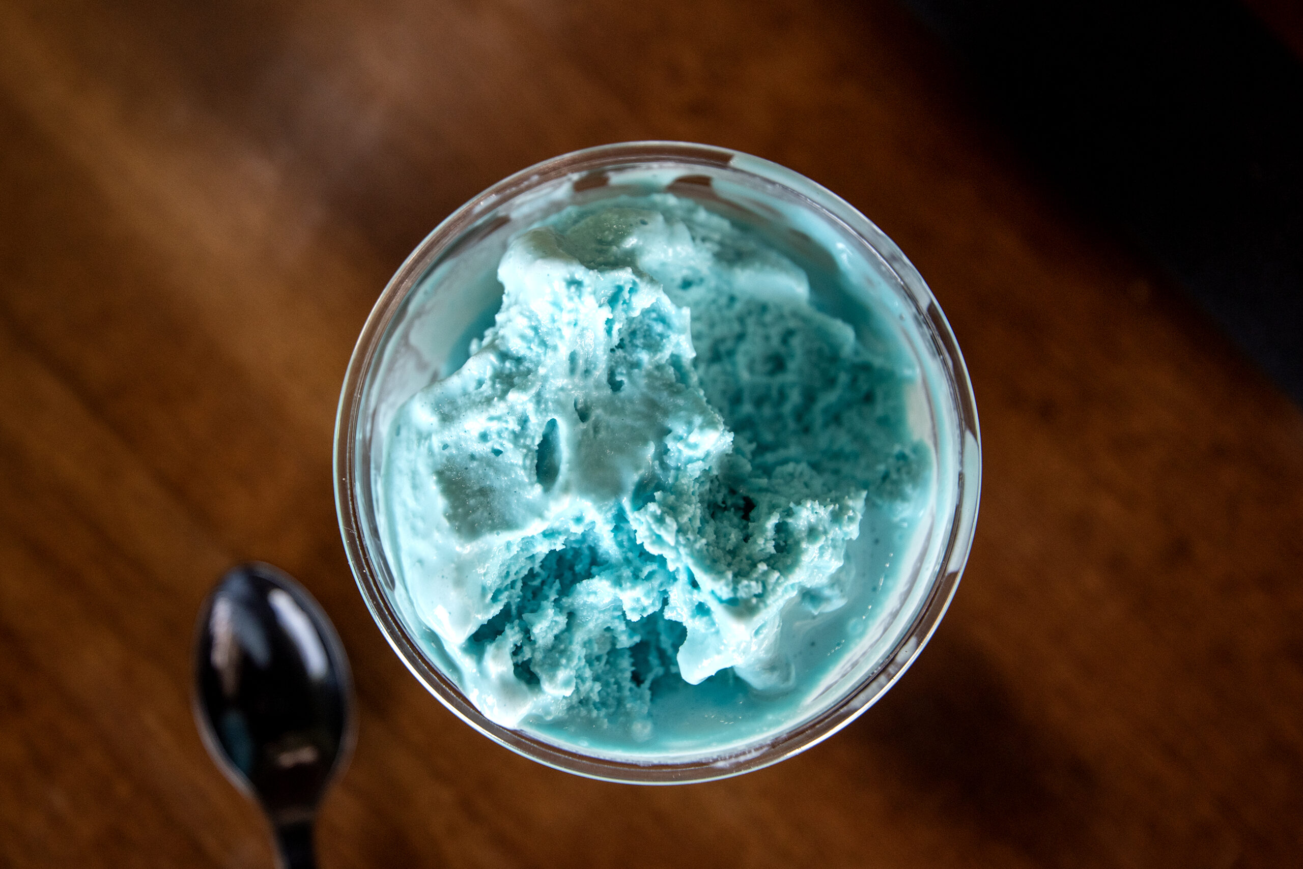 A glass cup filled with bright blue ice cream sits on a wooden surface next to a metal spoon.