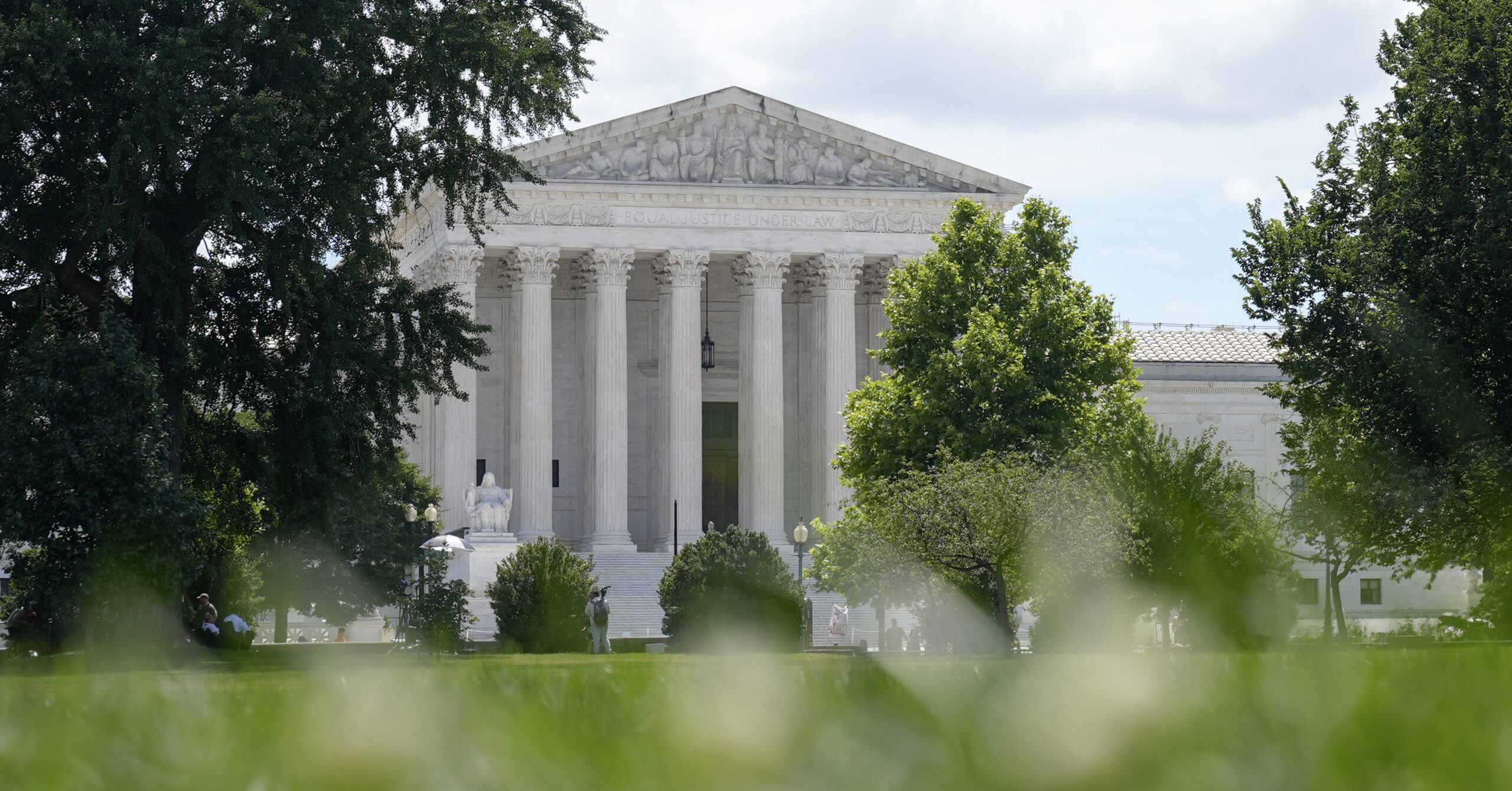 The United States Supreme Court building is seen in the background, partially obscured by green trees and grass in the foreground.